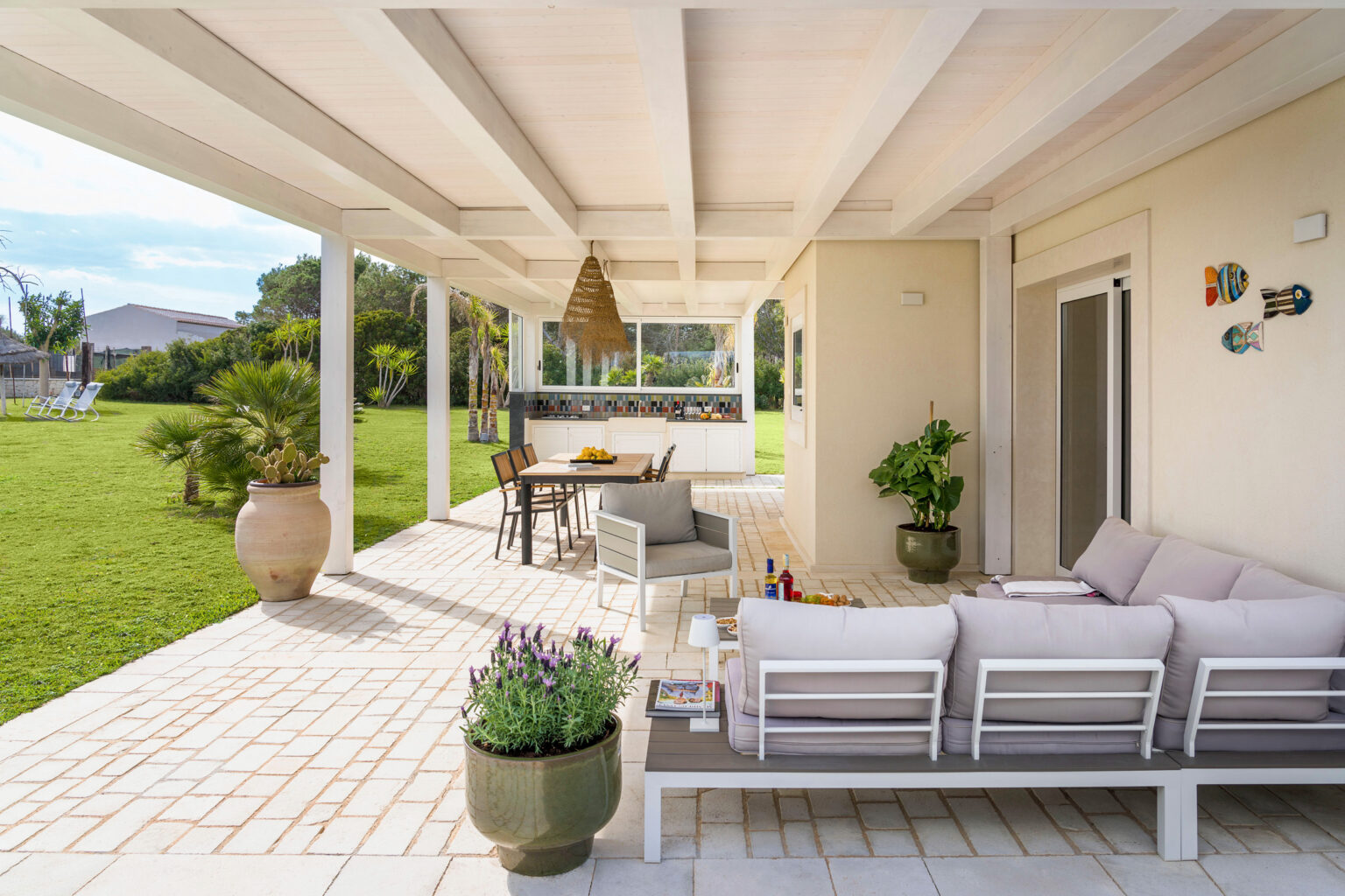 Modern outdoor patio featuring a stylish seating area with a plush sofa, a dining table, and an outdoor kitchen. Lush green lawn and tropical plants create a serene atmosphere, ideal for relaxation and entertaining. | Only on SopranoVillas