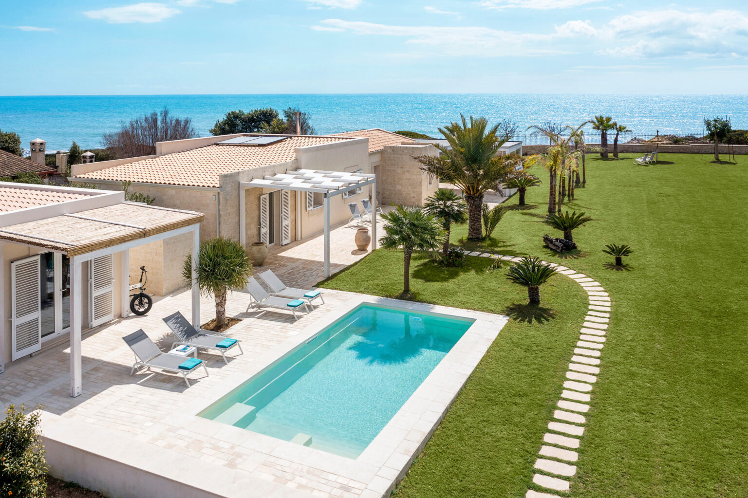Modern villa with a private pool and lush garden overlooking the ocean, featuring lounge chairs and palm trees, ideal for a relaxing getaway. | Only on SopranoVillas