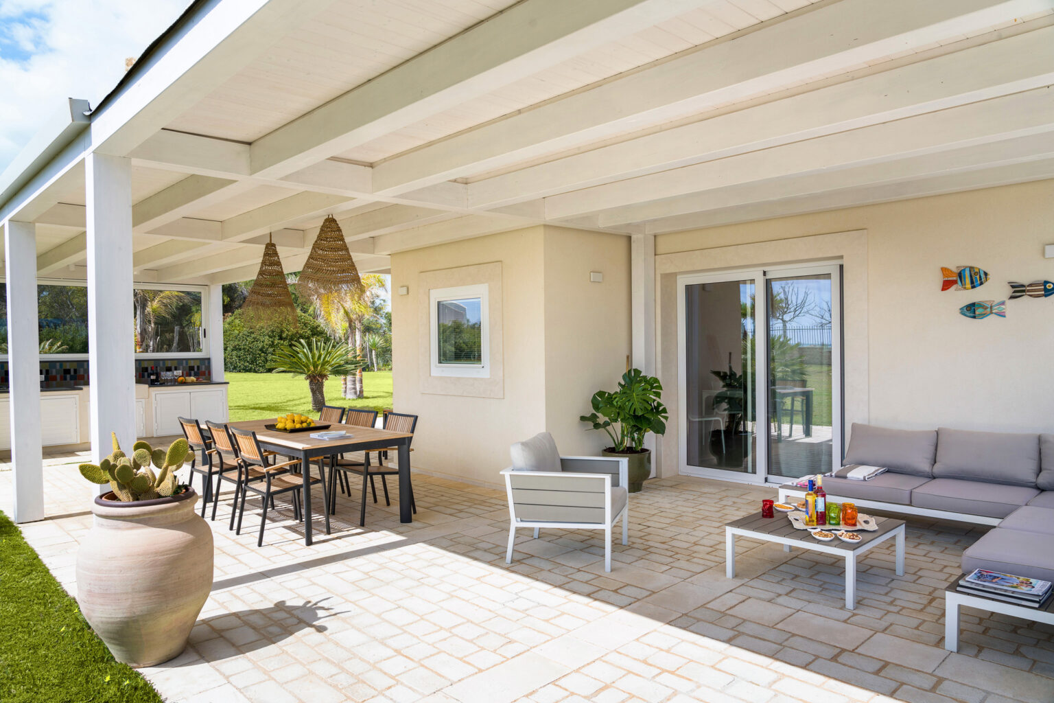 Modern outdoor patio with a dining table, comfortable seating, and decorative elements, featuring lush greenery and a view of the garden. The space is designed for relaxation and entertaining, with woven light fixtures and a stylish aesthetic. | Only on SopranoVillas