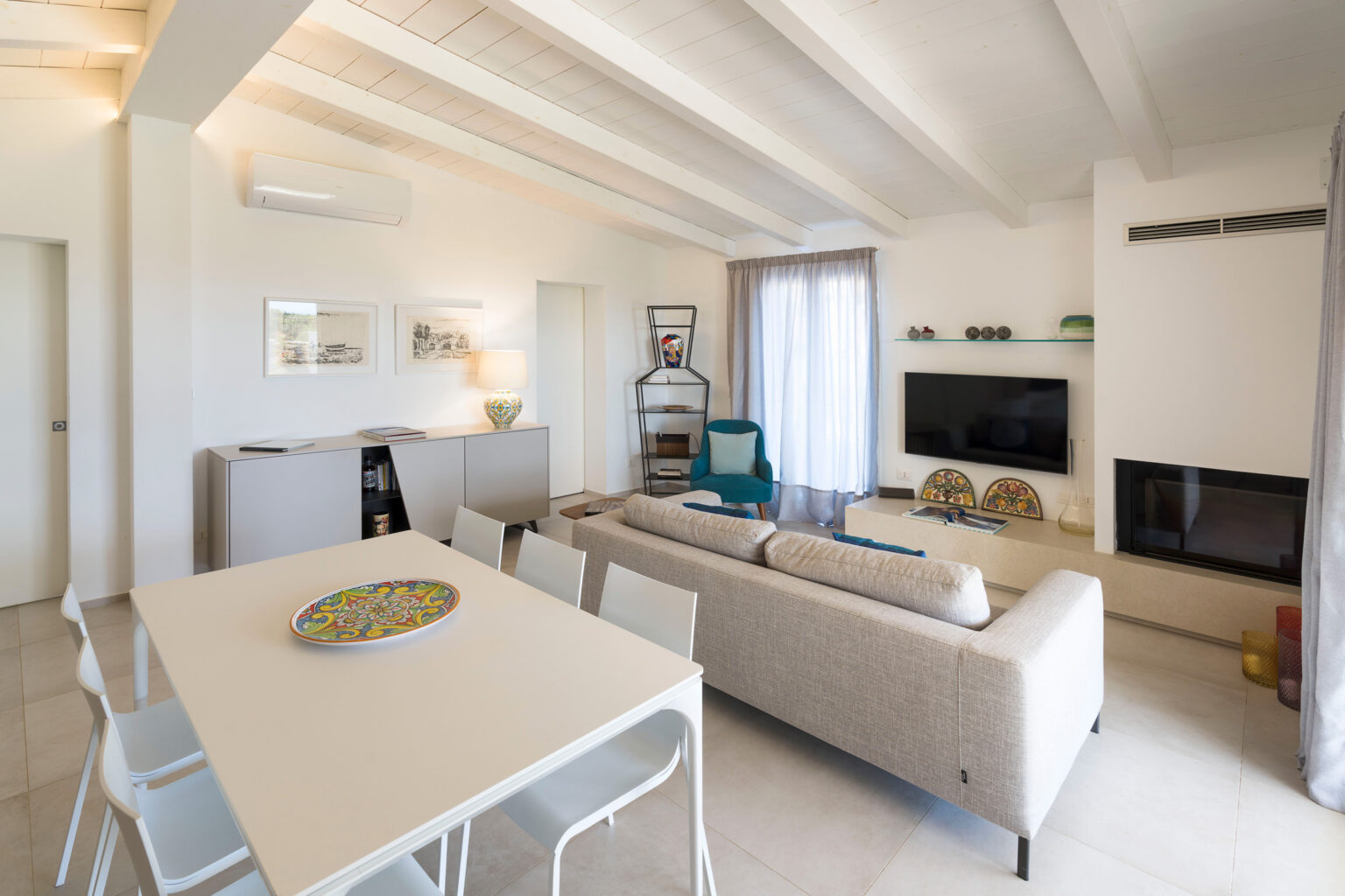 Modern living room with white walls, exposed wooden beams, a comfortable beige sofa, and a flat-screen TV. The space features a dining area with a white table and chairs, large windows with sheer curtains, and decorative elements that enhance the contemporary aesthetic. | Only on SopranoVillas