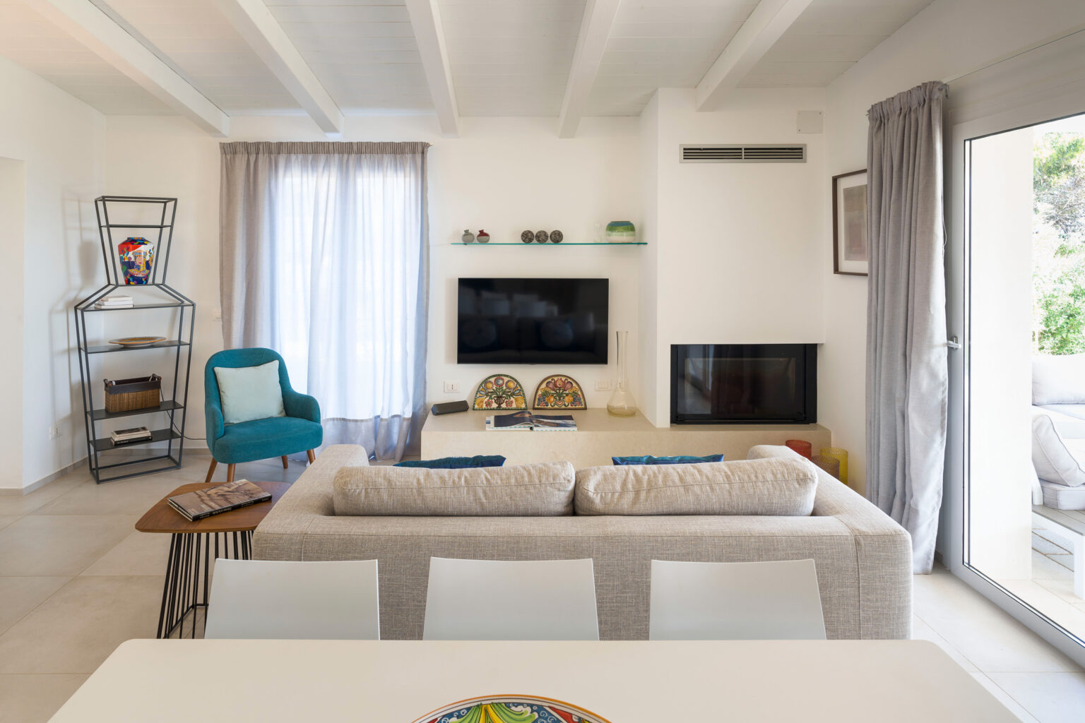 Modern living room interior featuring a neutral color palette, a comfortable beige sofa with blue cushions, a stylish turquoise armchair, and a contemporary shelving unit. The space includes a wall-mounted television, decorative items on a glass shelf, and large windows with sheer curtains allowing natural light. A dining table with white chairs is visible in the foreground, enhancing the open and airy feel of the room. | Only on SopranoVillas