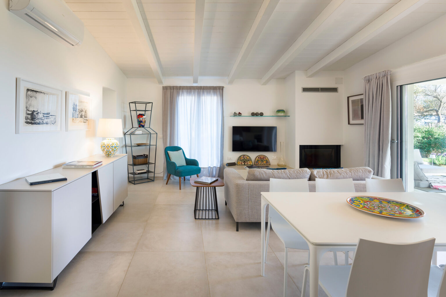 Modern living room and dining area featuring white walls, wooden beams, and stylish furniture. The space includes a comfortable sofa, a blue accent chair, a dining table with decorative tray, and large windows providing natural light. | Only on SopranoVillas