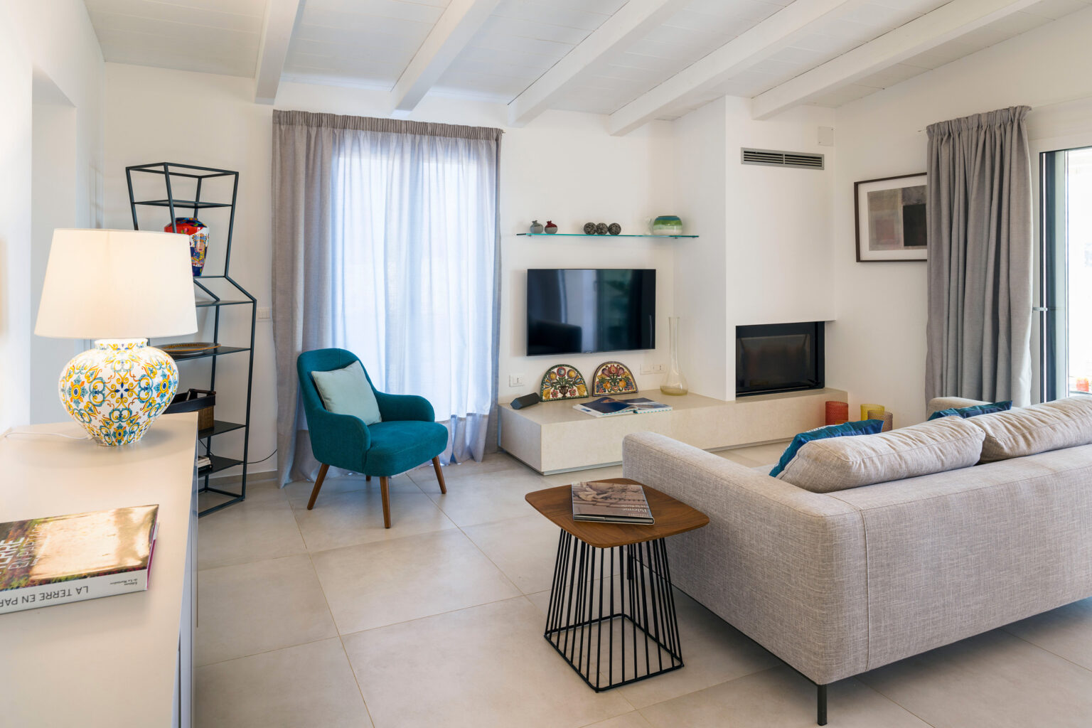 Modern living room featuring a stylish gray sofa, a blue accent chair, and a decorative lamp. The space includes a sleek TV stand, a cozy fireplace, and large windows with light curtains, creating a bright and inviting atmosphere. | Only on SopranoVillas
