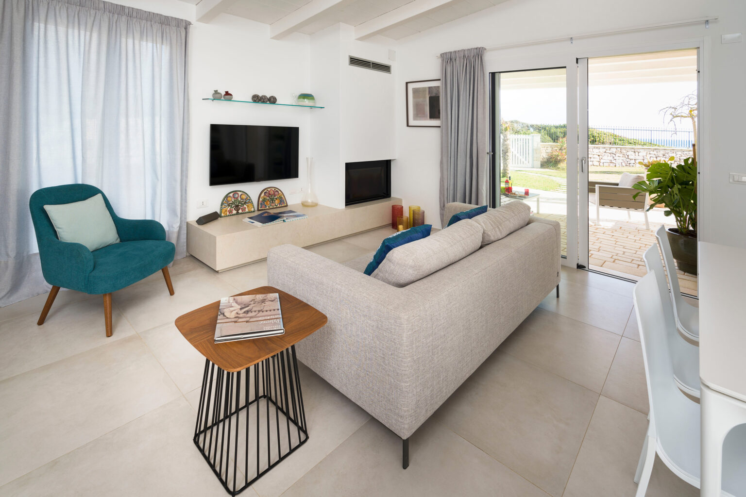 Modern living room featuring a light-colored sofa, a stylish blue accent chair, and a wooden coffee table. Large windows provide natural light and a view of the outdoor space. Minimalist decor includes a wall-mounted TV and decorative items on shelves. | Only on SopranoVillas