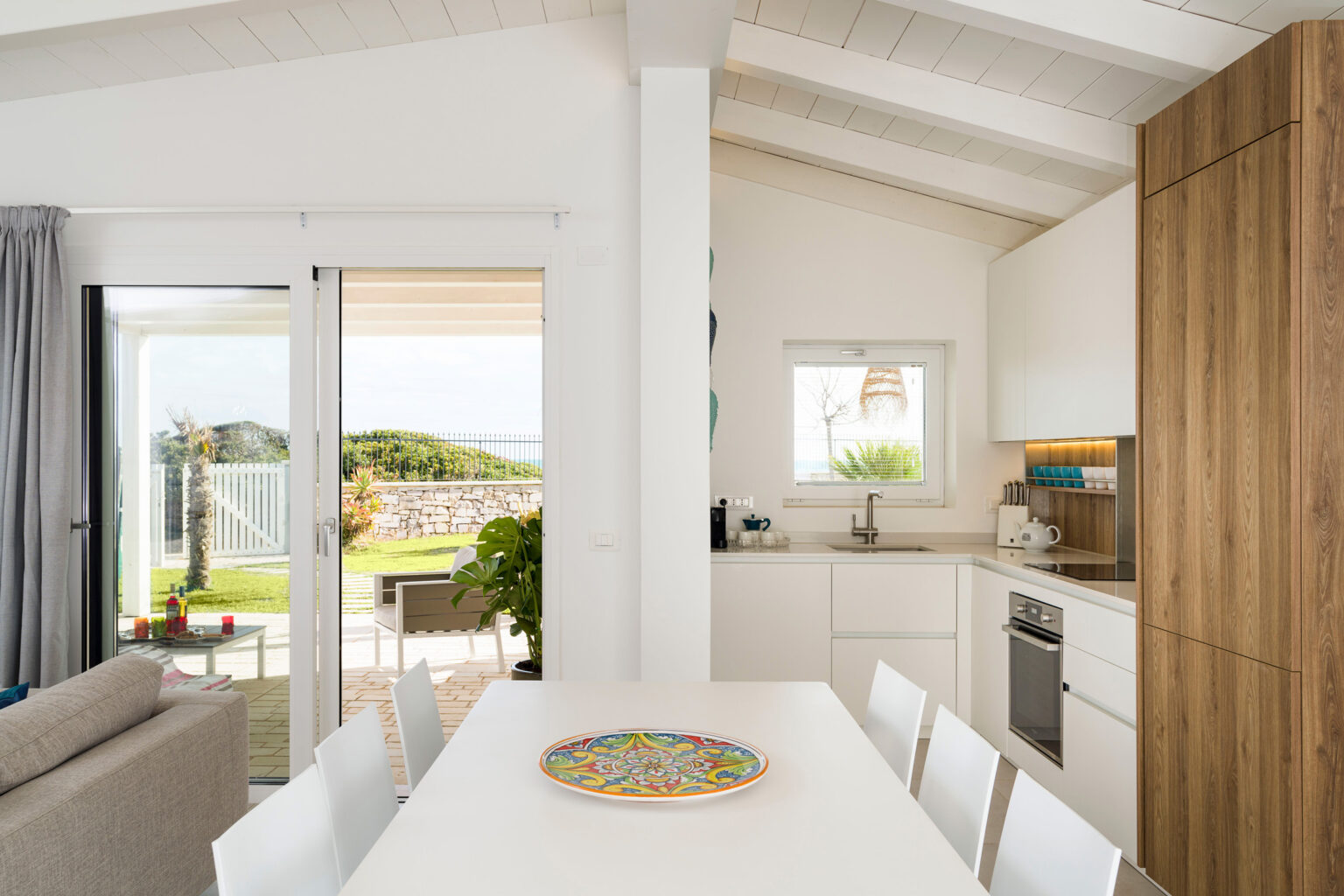 Modern kitchen and dining area in a bright, open-concept home with natural light, featuring a wooden dining table, white cabinetry, and sliding glass doors leading to a garden. | Only on SopranoVillas