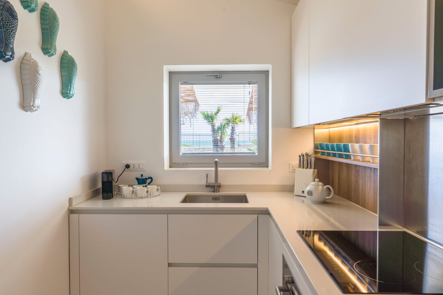 Modern kitchen interior featuring sleek white cabinetry, a stainless steel sink, and a window showcasing palm trees outside. The counter includes a coffee machine, a tray with teacups, and colorful dishware neatly arranged on a wooden shelf, enhancing the contemporary design. | Only on SopranoVillas