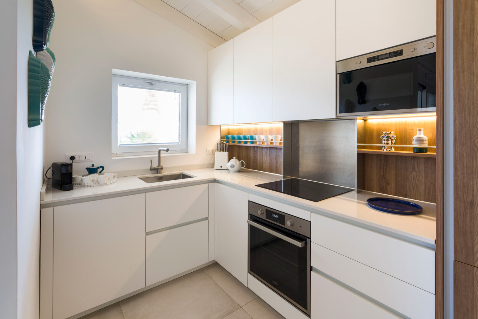 Modern kitchen featuring sleek white cabinetry, stainless steel appliances, and a minimalist design. The space includes a sink, stove, and ample countertop area, illuminated by under-cabinet lighting, with a window providing natural light. | Only on SopranoVillas