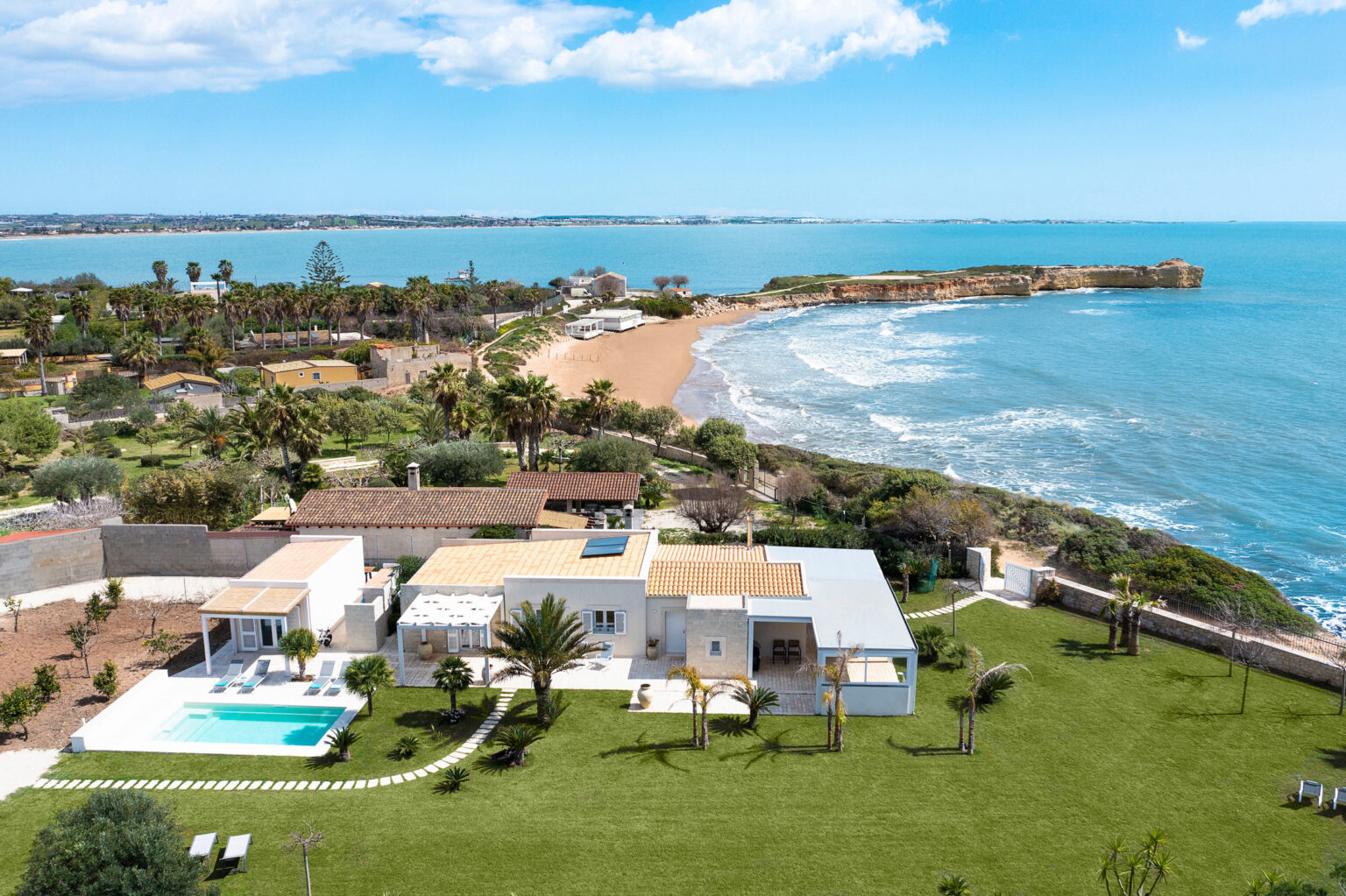Aerial view of a modern coastal villa with a swimming pool, surrounded by lush greenery and overlooking a sandy beach and the ocean in the background. | Only on SopranoVillas