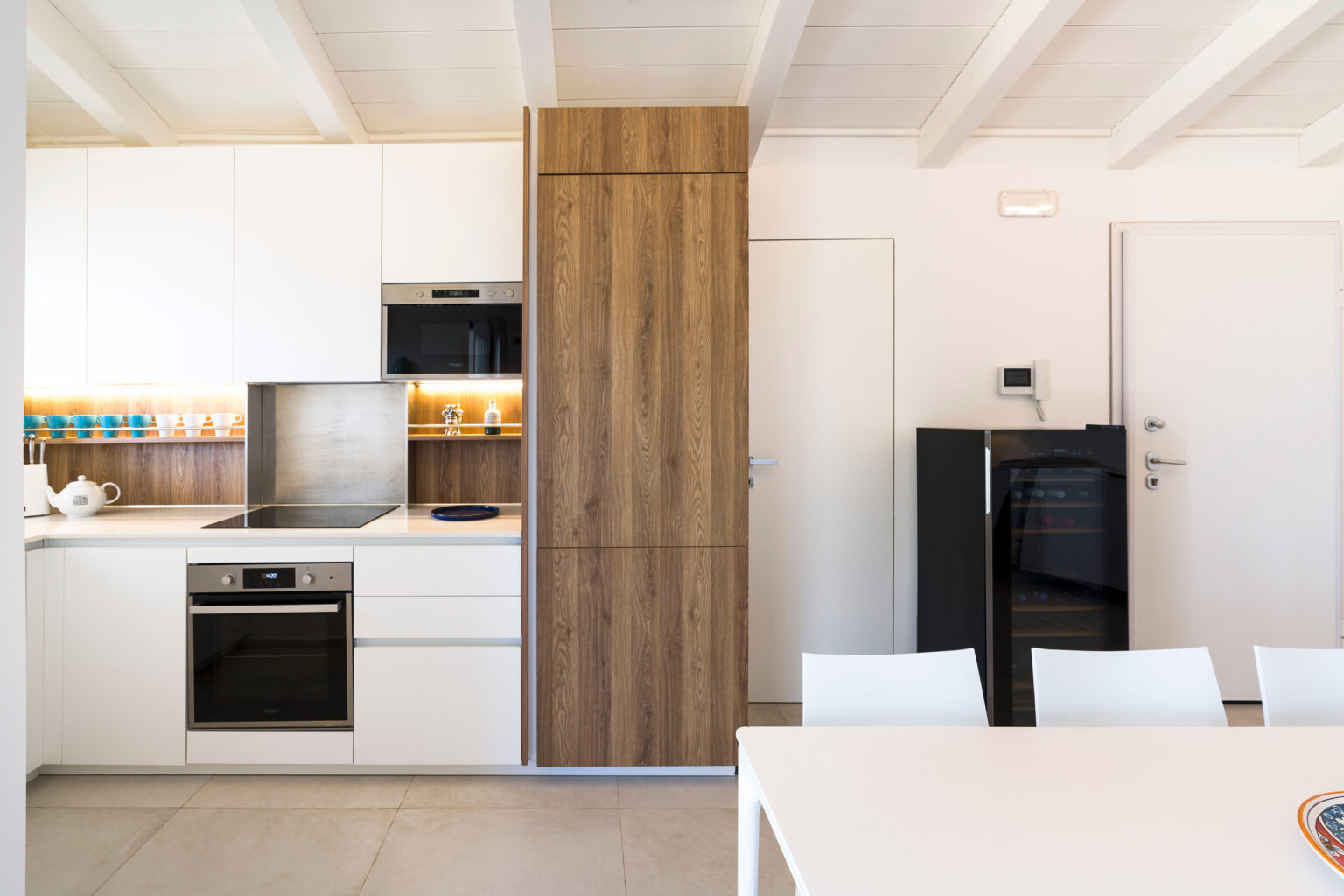 Modern kitchen interior featuring sleek white cabinetry, a wooden accent refrigerator, built-in oven, and a dining table with white chairs. The space is well-lit with natural light, showcasing a minimalist design. | Only on SopranoVillas
