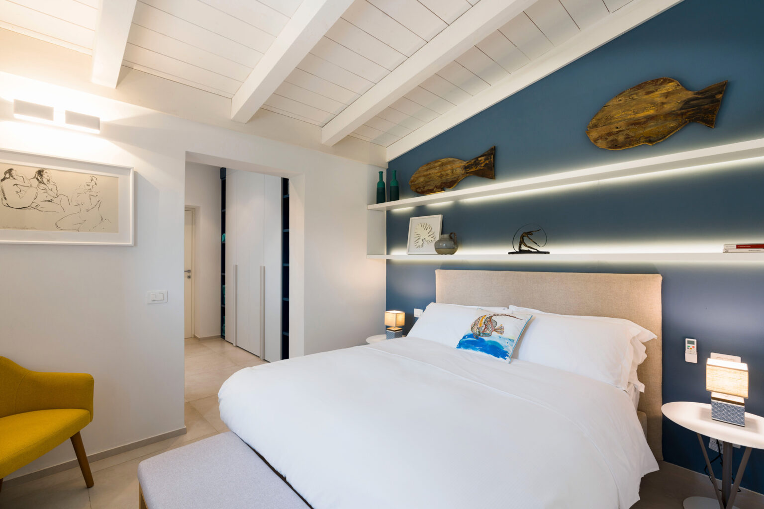 Modern bedroom design featuring a cozy bed with white bedding, a yellow accent chair, and decorative fish wall art against a blue wall. The room includes stylish lighting and minimalist decor, creating a serene atmosphere. | Only on SopranoVillas