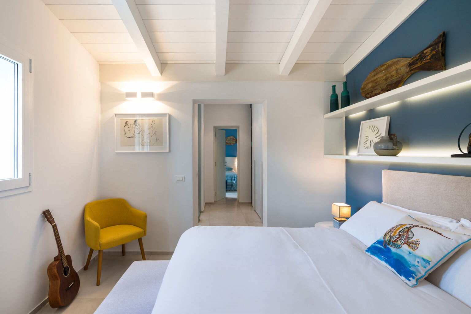 Modern bedroom with white walls and wooden beams, featuring a comfortable bed, decorative pillows, an accent chair, and a guitar. The room has a blue accent wall and stylish shelving with decor, creating a cozy and inviting atmosphere. | Only on SopranoVillas