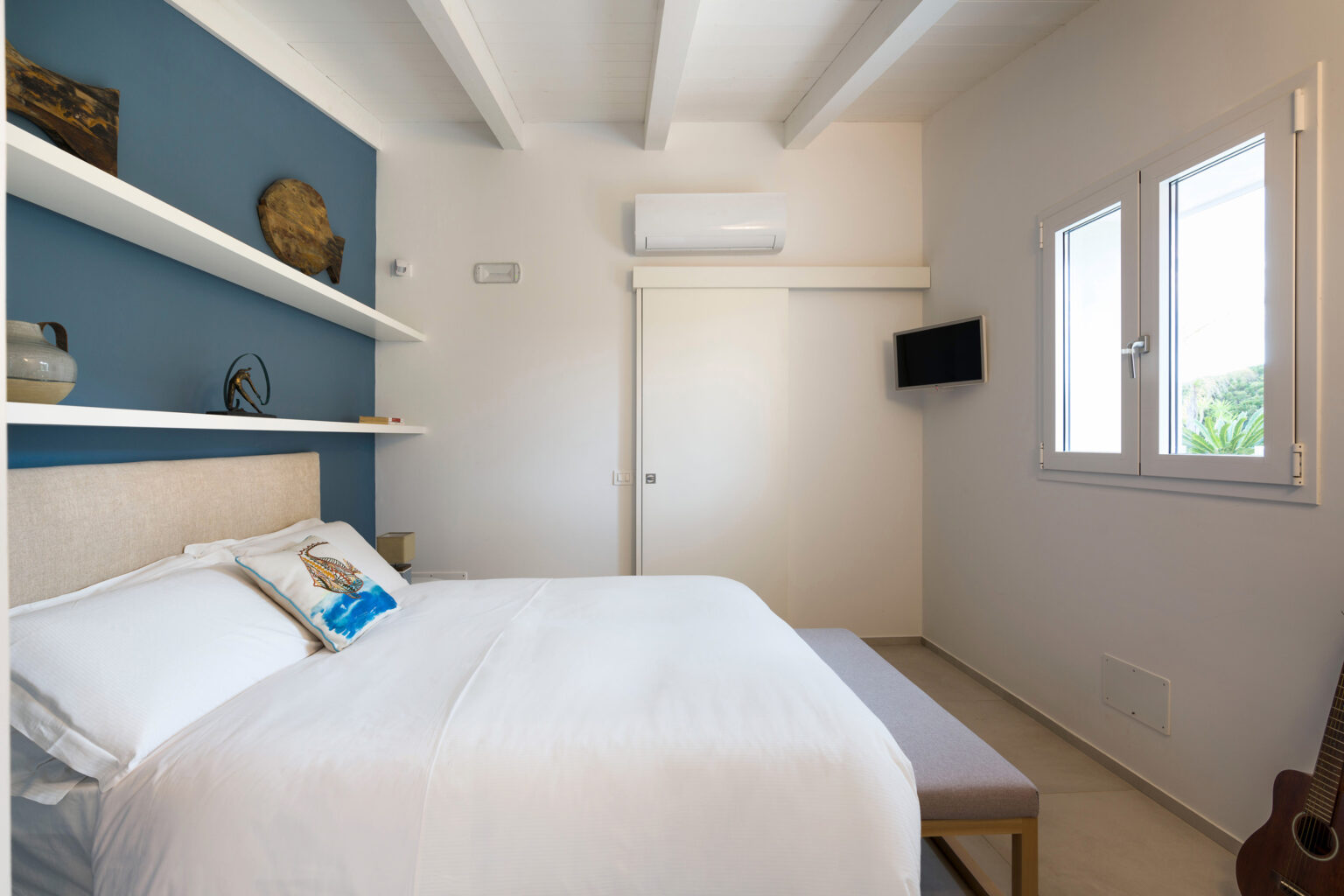 Modern bedroom featuring a comfortable queen-size bed, minimalist decor, and a soothing blue accent wall. The room includes shelves with decorative items, a wall-mounted TV, and ample natural light from large windows. Ideal for relaxation and comfort. | Only on SopranoVillas