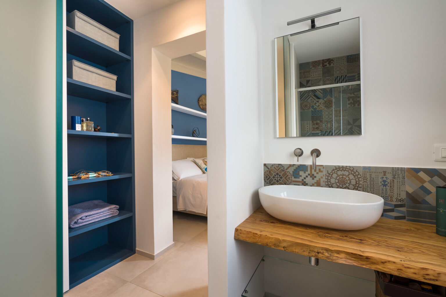 Modern bathroom design featuring a stylish sink on a wooden countertop, a large mirror, and blue shelving for storage. The space is bright and airy, with neutral tones and a glimpse of a bedroom in the background. | Only on SopranoVillas
