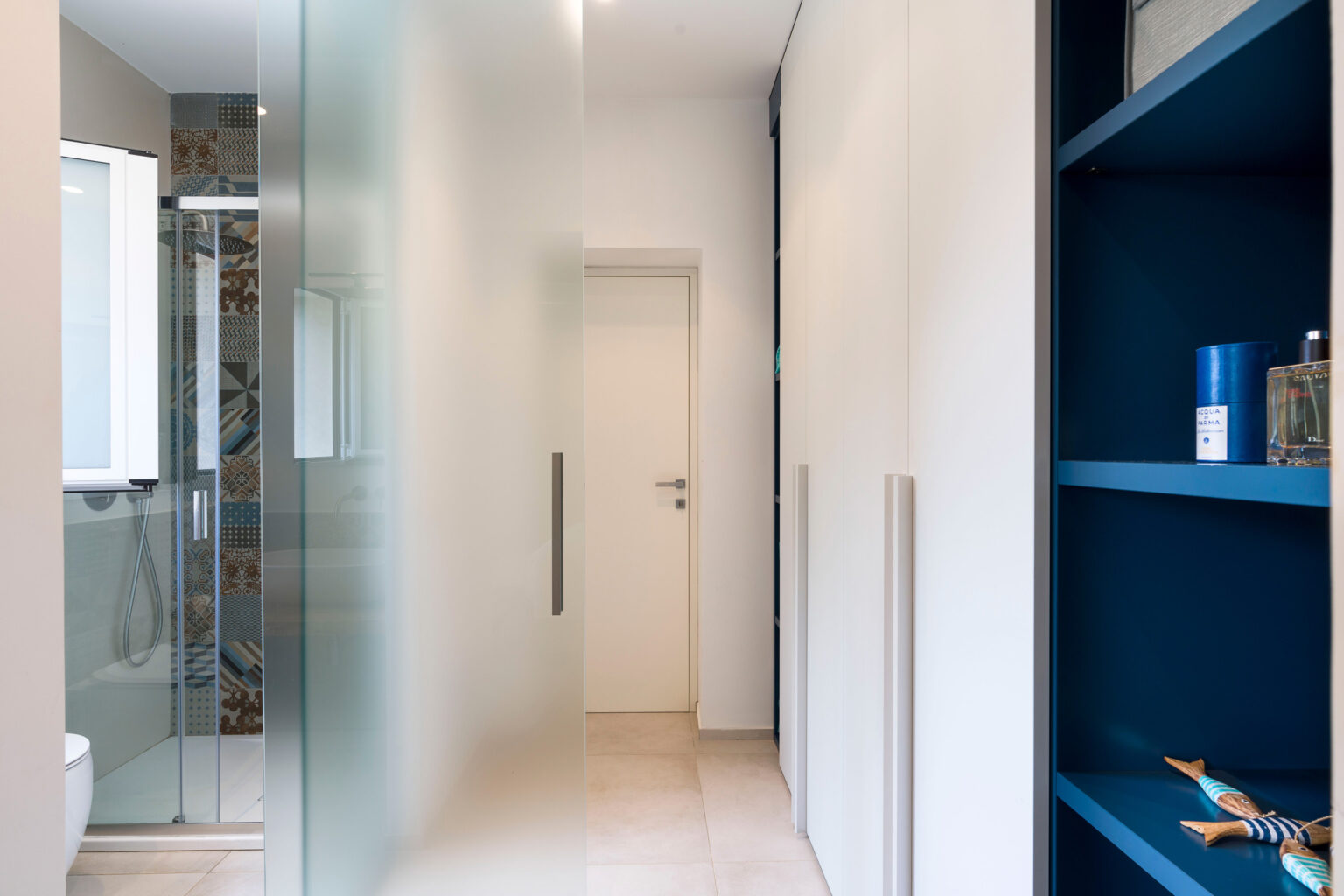 Modern hallway featuring a frosted glass door, minimalist design, and a blue shelving unit with decorative items. The light-colored walls and tiled floor create a spacious and inviting atmosphere. | Only on SopranoVillas