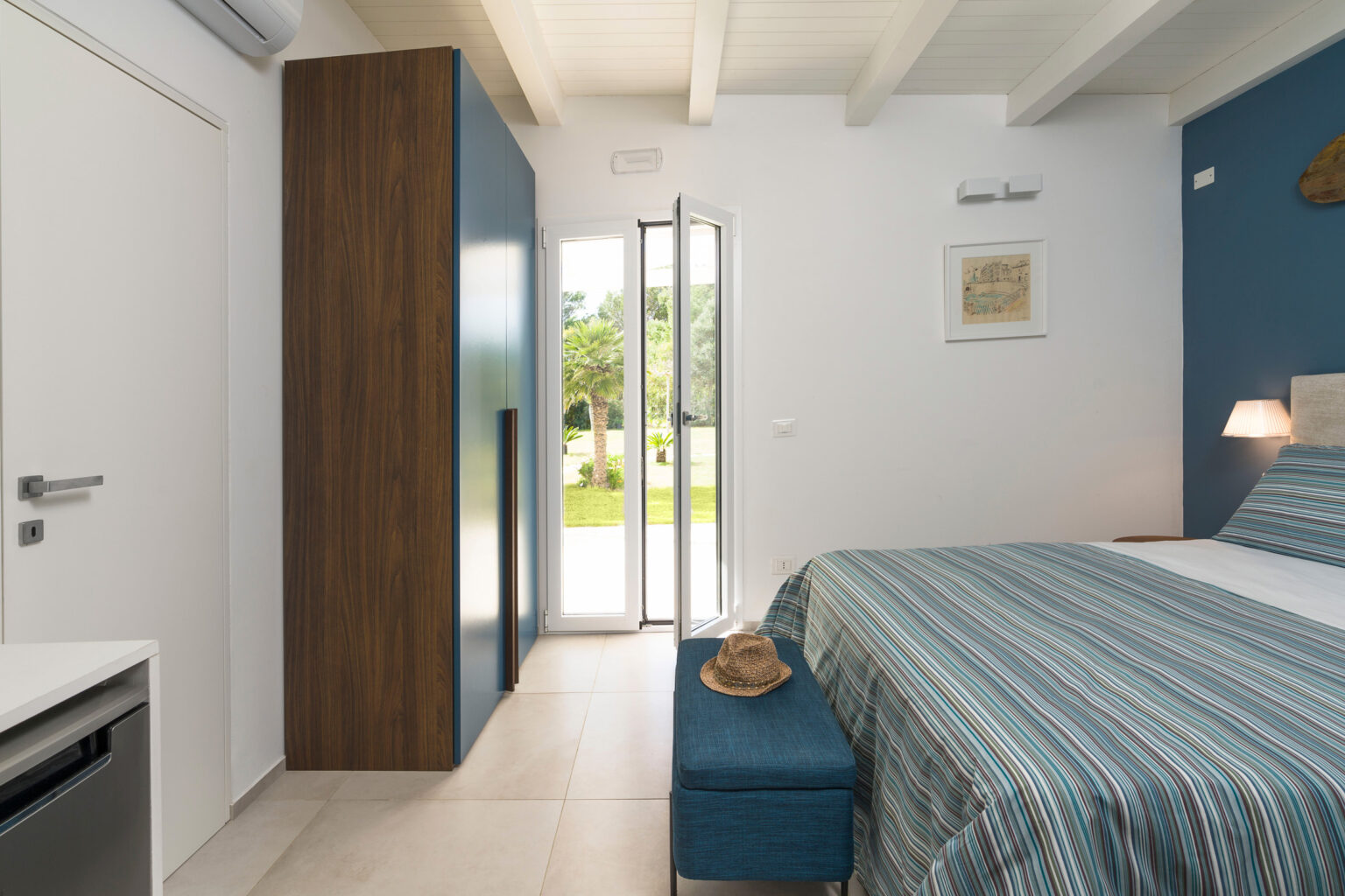 Modern bedroom with blue accent wall, featuring a striped bedspread, bedside lamp, and a doorway leading to an outdoor view. Includes a wardrobe and a small seating area. Ideal for relaxation and comfort. | Only on SopranoVillas