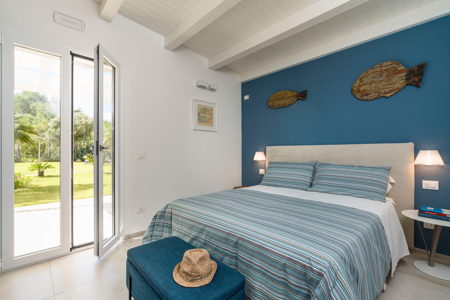 Cozy bedroom featuring a blue accent wall, a comfortable bed with striped bedding, two bedside lamps, and large glass doors opening to a scenic garden view. Decorated with fish wall art and a stylish hat on a bench, the room exudes a relaxing coastal vibe. | Only on SopranoVillas