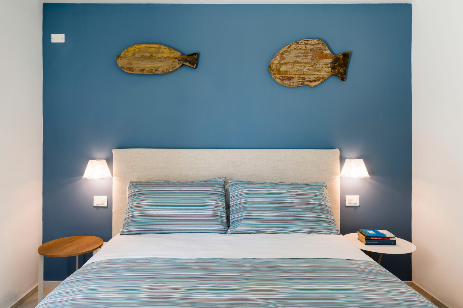 Cozy bedroom featuring a light blue accent wall with wooden fish decor, a neatly made bed with striped blue and white bedding, and modern bedside lamps on either side. | Only on SopranoVillas
