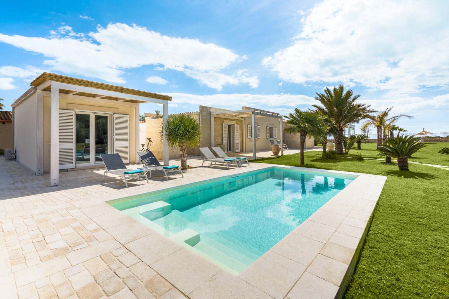 Modern villa with a private pool, surrounded by lush greenery and palm trees, featuring outdoor lounge chairs and a clear blue sky. Ideal for relaxation and luxury vacationing. | Only on SopranoVillas