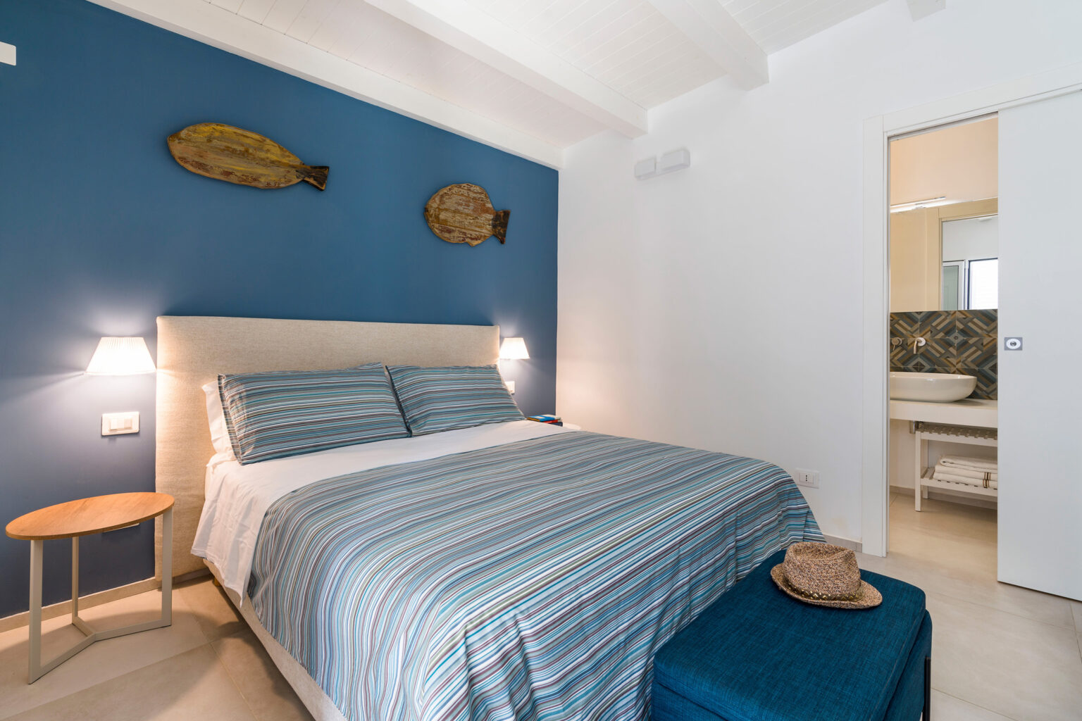 Cozy bedroom featuring a striped bedspread, modern lighting, and decorative fish wall art. The room includes a bedside table and a teal ottoman, with an open door leading to a stylish bathroom. | Only on SopranoVillas