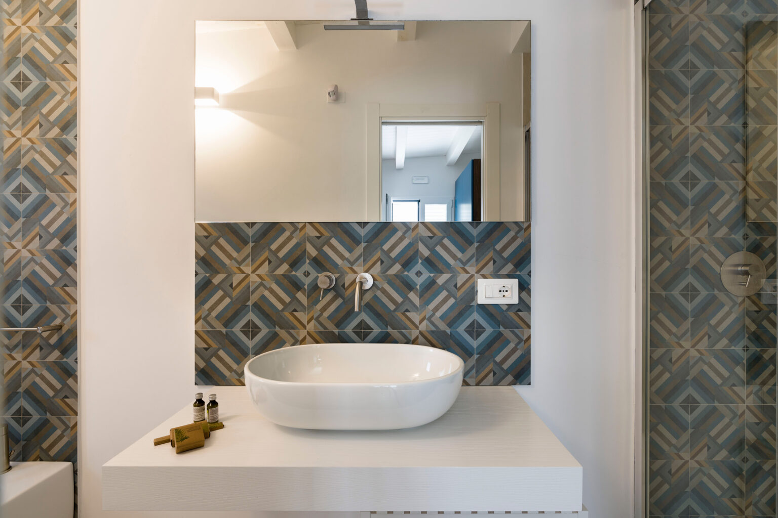 Modern bathroom featuring a stylish white vessel sink on a minimalist countertop, with patterned blue and gold wall tiles creating an elegant backdrop. Natural light enhances the contemporary design, making it a serene space. | Only on SopranoVillas