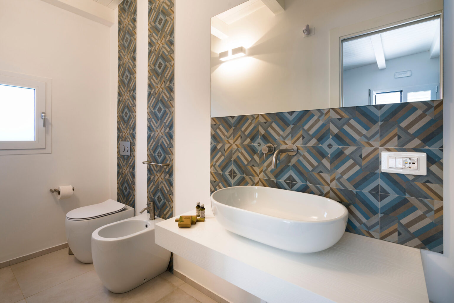 Modern bathroom featuring a stylish design with patterned blue and brown tiles, a white oval sink on a floating shelf, a bidet, and a toilet. The space is illuminated by wall-mounted lighting and includes a mirror and a window for natural light. | Only on SopranoVillas