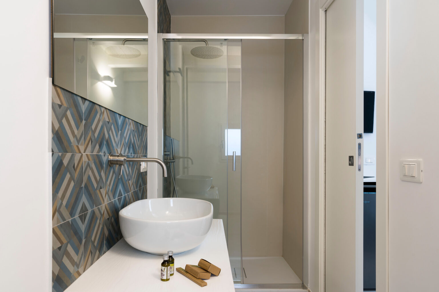 Modern bathroom interior featuring a stylish glass shower, a sleek white sink, and decorative patterned tiles. Natural light enhances the space, complemented by essential oils and towels on the counter. Ideal for showcasing contemporary bathroom design. | Only on SopranoVillas