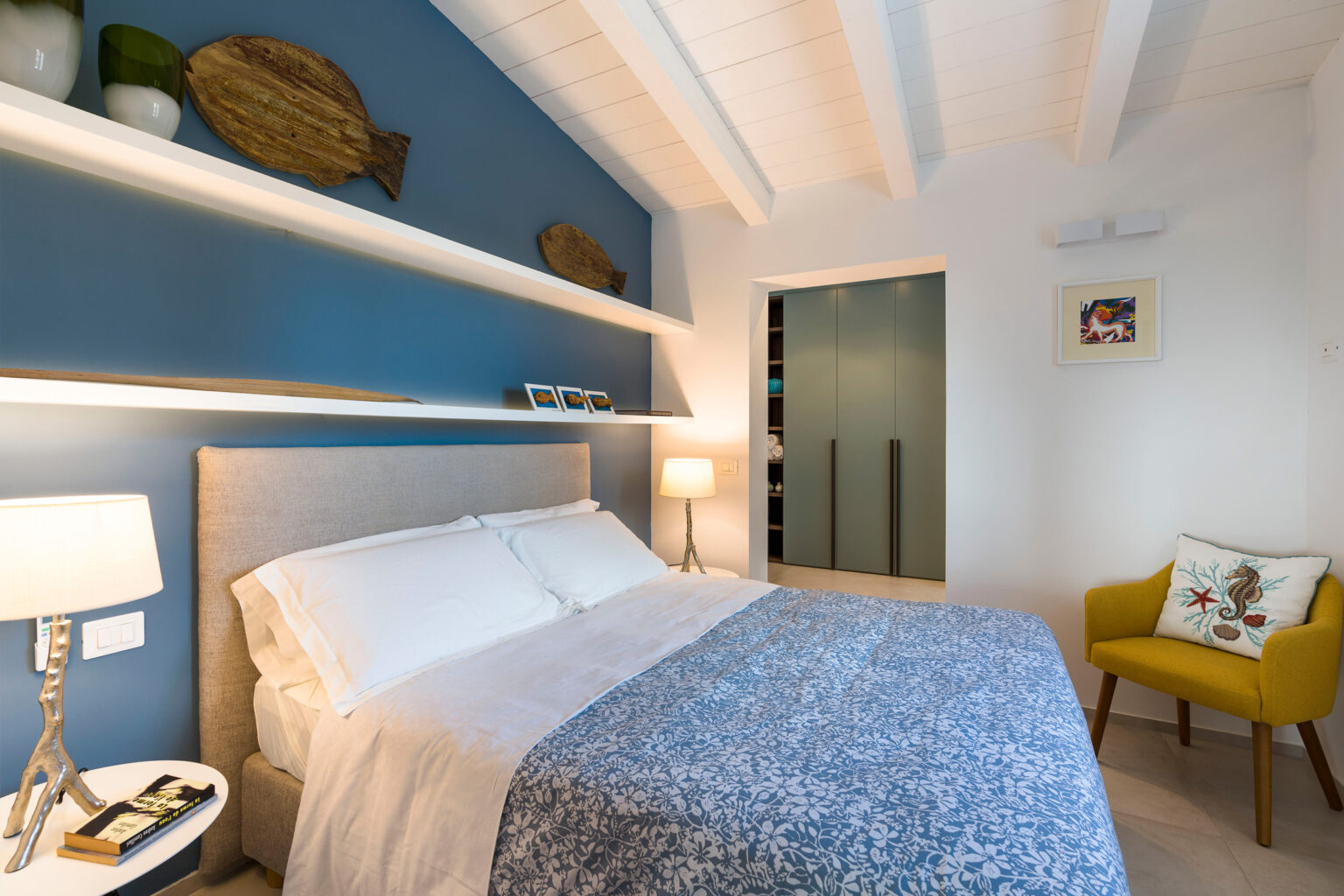 Cozy modern bedroom featuring a plush bed with white linens and a blue patterned throw blanket, complemented by stylish decor including fish-shaped wall art and a vibrant yellow chair. The room showcases a minimalist design with white wooden beams and built-in storage, creating a serene and inviting atmosphere. Ideal for relaxation and comfort. | Only on SopranoVillas