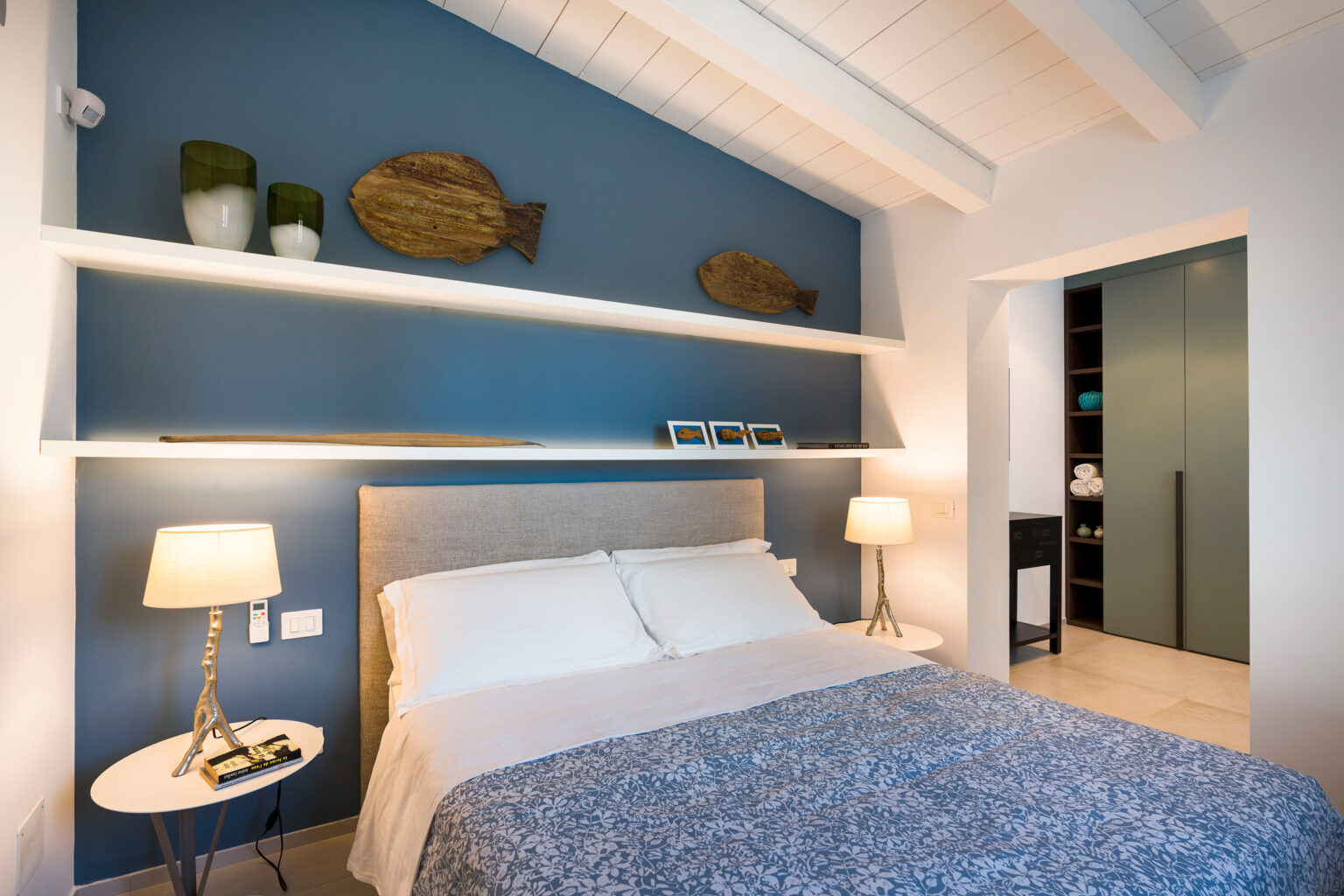 Cozy bedroom featuring a light gray upholstered bed with white linens and a blue floral throw blanket, accented by a modern bedside lamp. The wall behind the bed is painted in a calming blue, adorned with wooden fish decor and stylish shelves displaying decorative vases and framed photos. A doorway leads to a closet with neatly arranged towels, enhancing the room's serene atmosphere. | Only on SopranoVillas