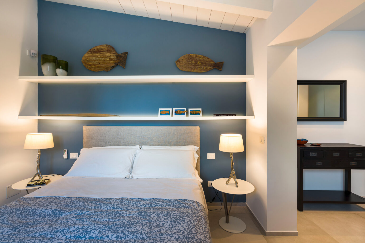 Cozy bedroom interior featuring a blue accent wall, a queen-sized bed with white linens, decorative fish wall art, and modern bedside lamps. The room includes a stylish black dresser and a mirror, creating a serene and inviting atmosphere. | Only on SopranoVillas