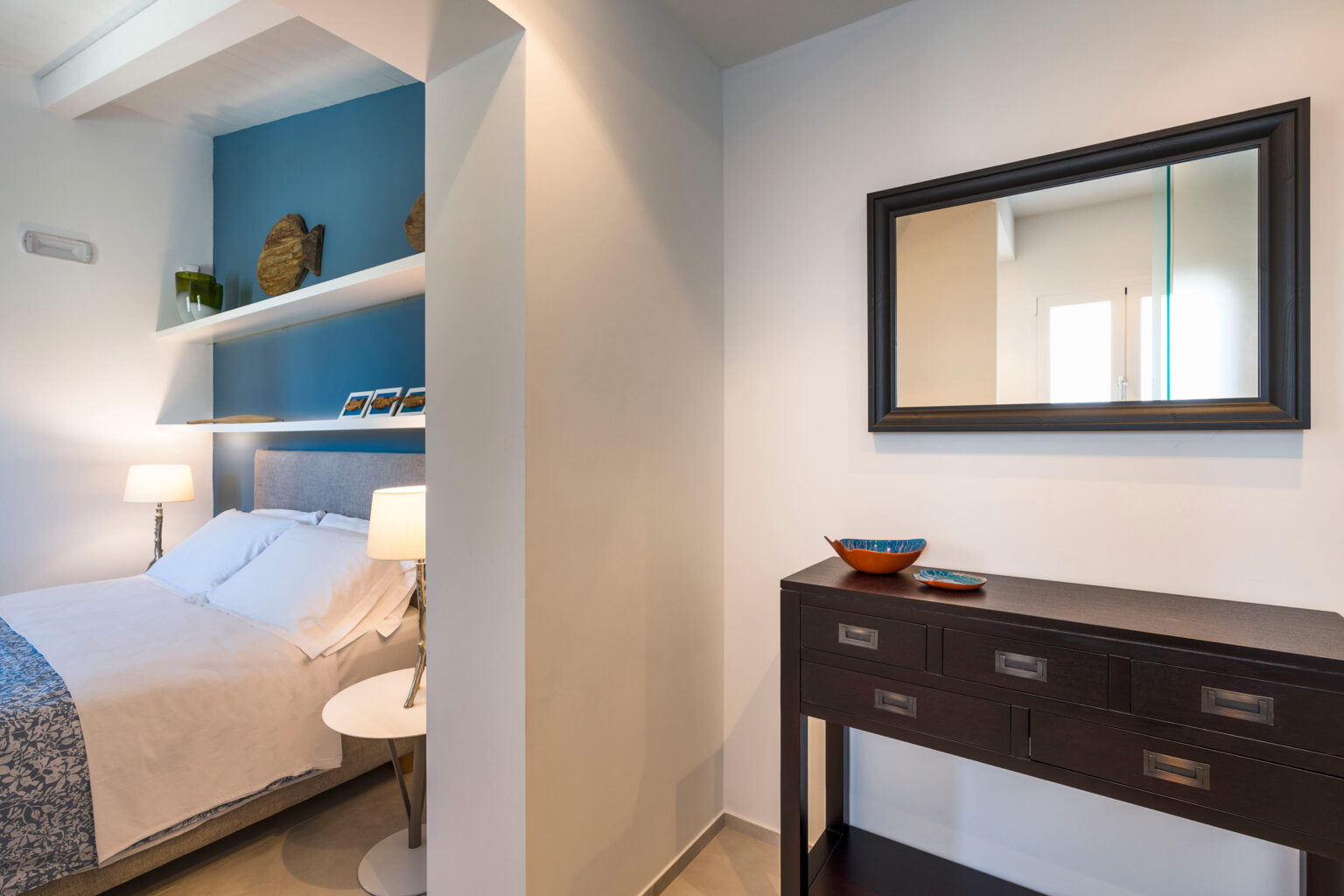 Modern bedroom interior featuring a cozy bed with white linens, blue accent wall, stylish mirror, and a dark wood console table with decorative bowl. Ideal for showcasing contemporary home decor. | Only on SopranoVillas