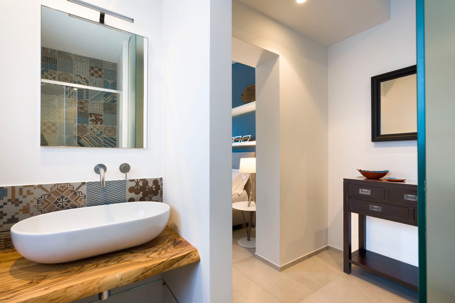 Modern bathroom interior featuring a sleek white vessel sink on a wooden countertop, decorative tiled backsplash, and a large mirror above. The space opens to a cozy nook with a lamp and decorative items, showcasing a blend of contemporary design and functional layout. | Only on SopranoVillas