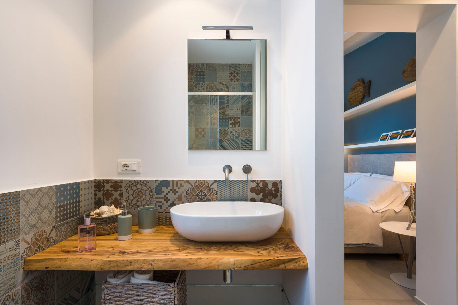 Modern bathroom with a wooden countertop, a round white sink, and a large mirror above. The wall features decorative tiles, and a glimpse of a bedroom with blue walls is visible in the background. | Only on SopranoVillas