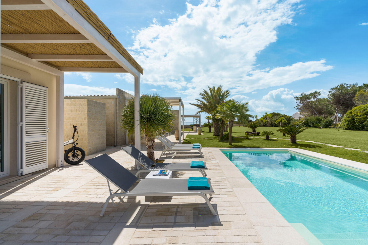 Luxurious outdoor pool area featuring modern lounge chairs, vibrant palm trees, and a serene landscape under a bright blue sky. Ideal for relaxation and leisure activities. | Only on SopranoVillas