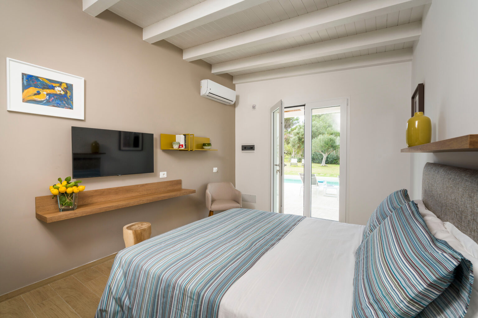 Cozy bedroom featuring a striped bedspread, a wall-mounted TV, and a modern decor with natural light streaming in through glass doors leading to a pool area. | Only on SopranoVillas