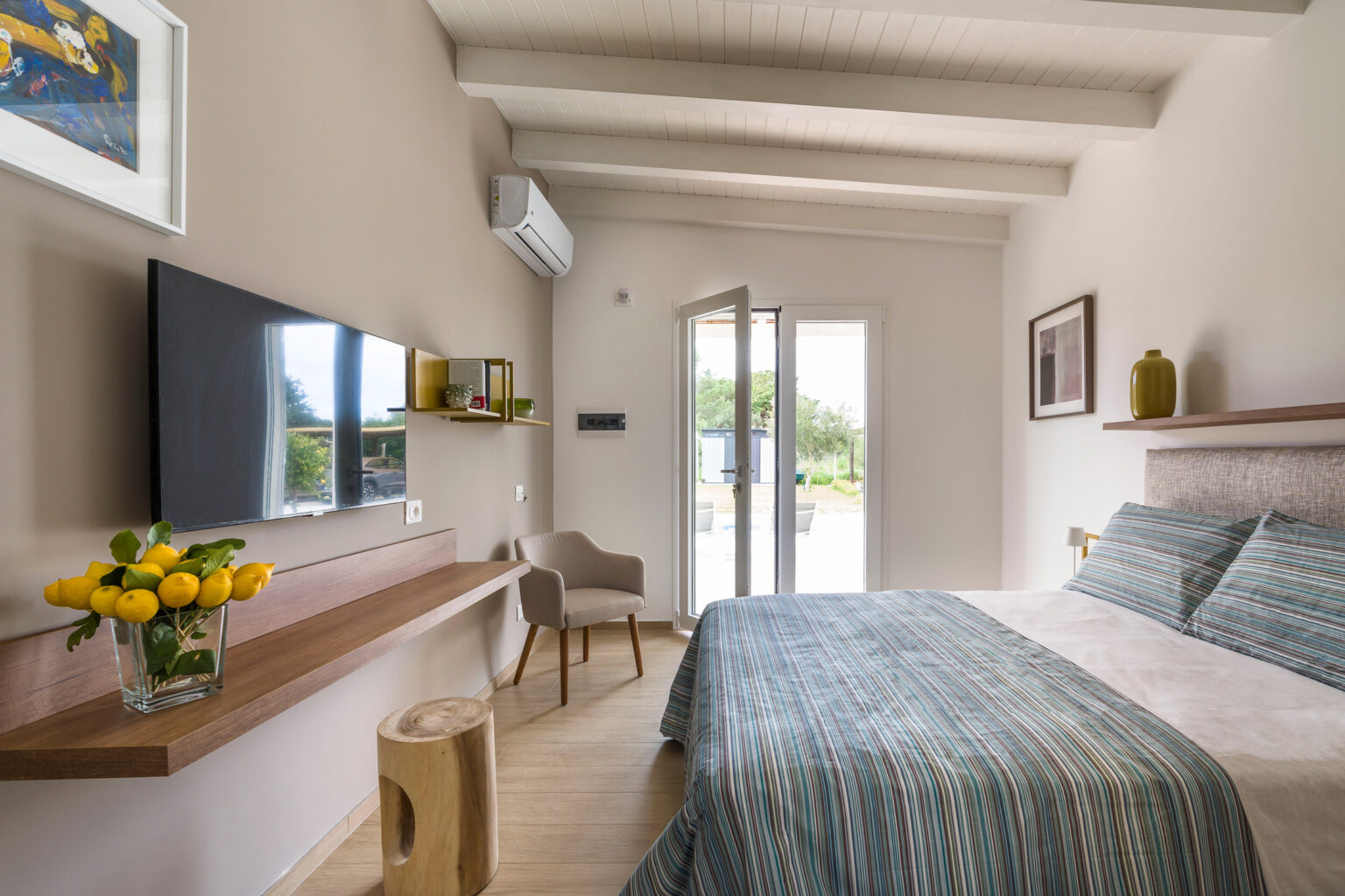 Cozy modern bedroom featuring a striped bedspread, wooden accents, and a flat-screen TV, with natural light streaming in from a glass door that opens to an outdoor area. The room includes a chair, decorative vase with lemons, and air conditioning for comfort. | Only on SopranoVillas