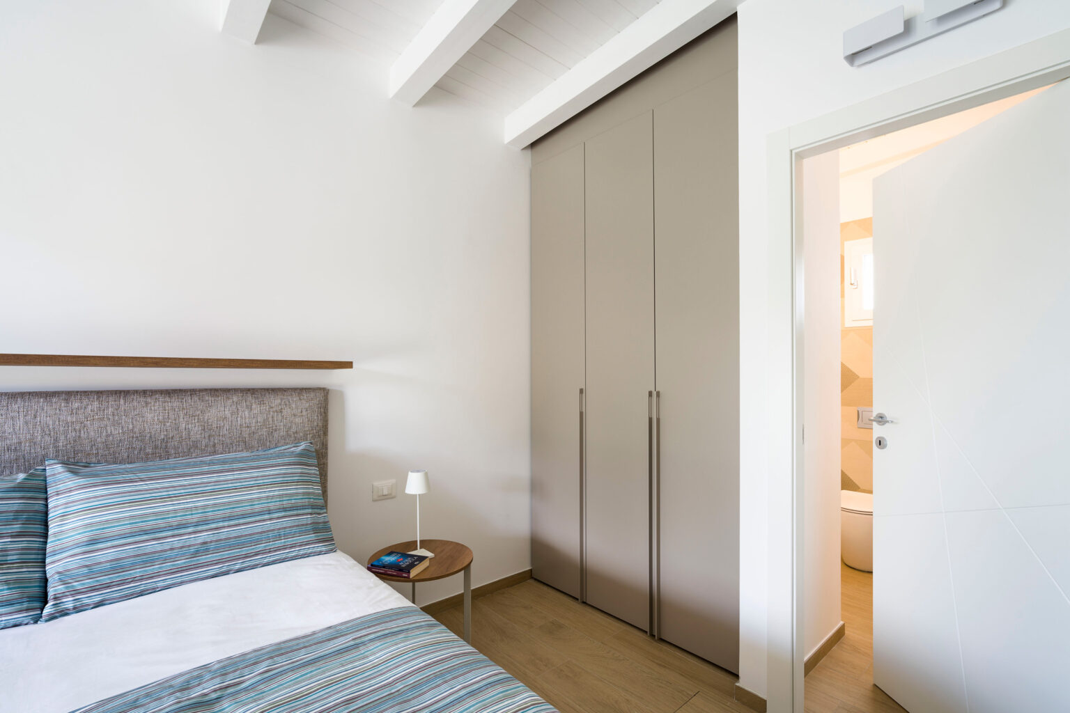 Modern bedroom interior featuring a neatly made bed with striped bedding, a small nightstand with a lamp, and a sliding closet door. The room has white walls and exposed beams, with a doorway leading to a bathroom. | Only on SopranoVillas
