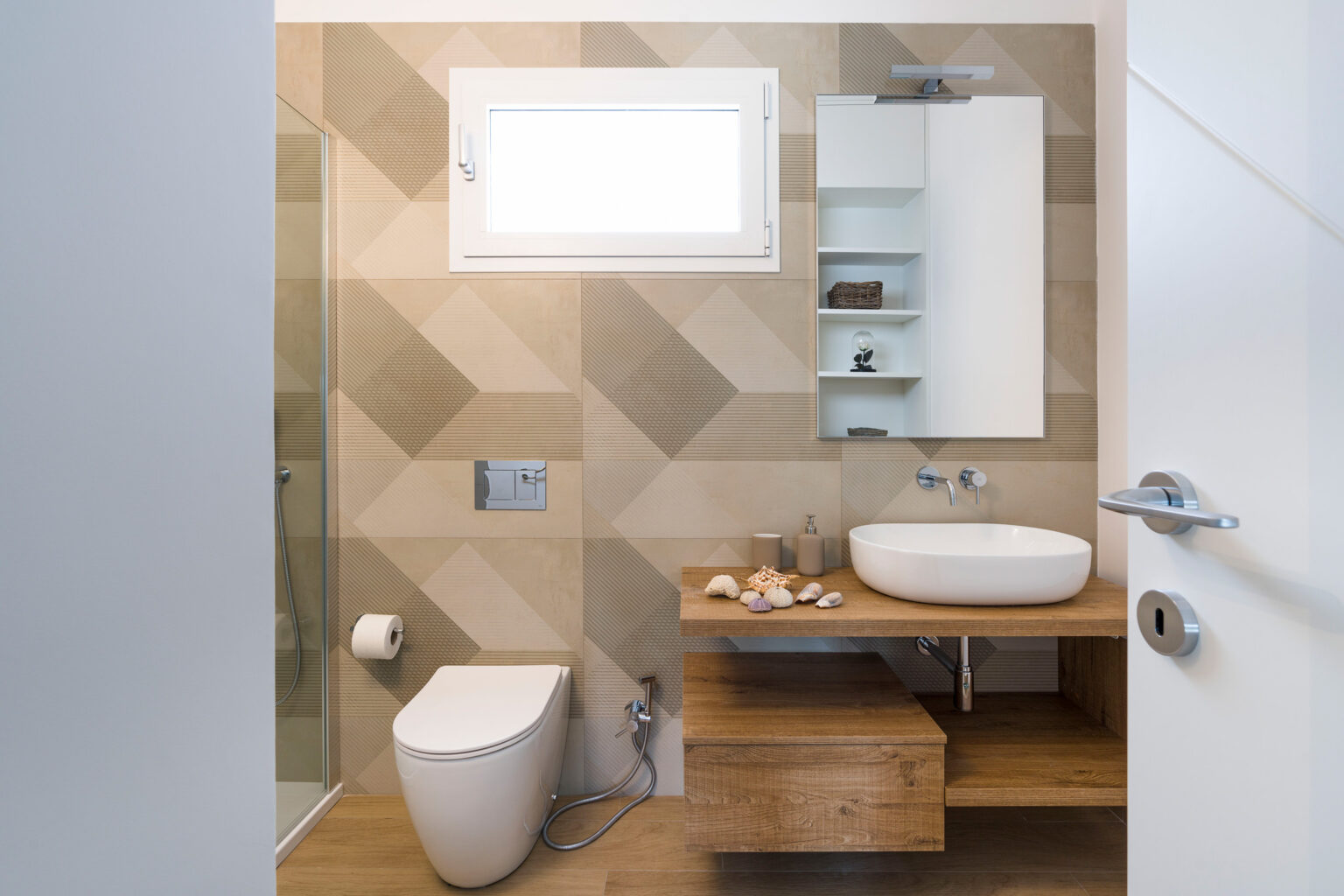 Modern bathroom design featuring a stylish wall with geometric tiles, a wooden vanity with a round sink, and a glass shower enclosure. The space is well-lit with natural light from a window and includes minimalistic storage solutions. | Only on SopranoVillas