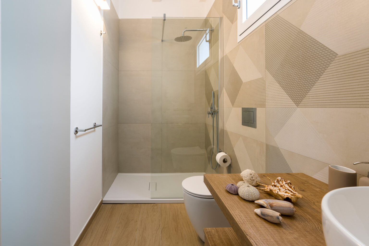Modern bathroom featuring a glass shower enclosure, neutral-toned geometric wall tiles, a wooden countertop with decorative shells, and a sleek toilet. Natural light enters through a window, enhancing the contemporary design. | Only on SopranoVillas