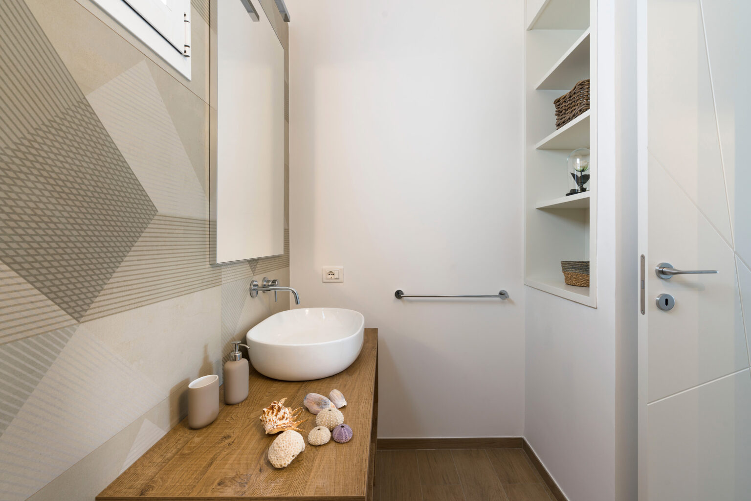 Modern bathroom interior featuring a stylish round sink on a wooden countertop, decorative seashells, and a minimalist shelving unit. The walls display a contemporary geometric design, enhancing the overall aesthetic. | Only on SopranoVillas