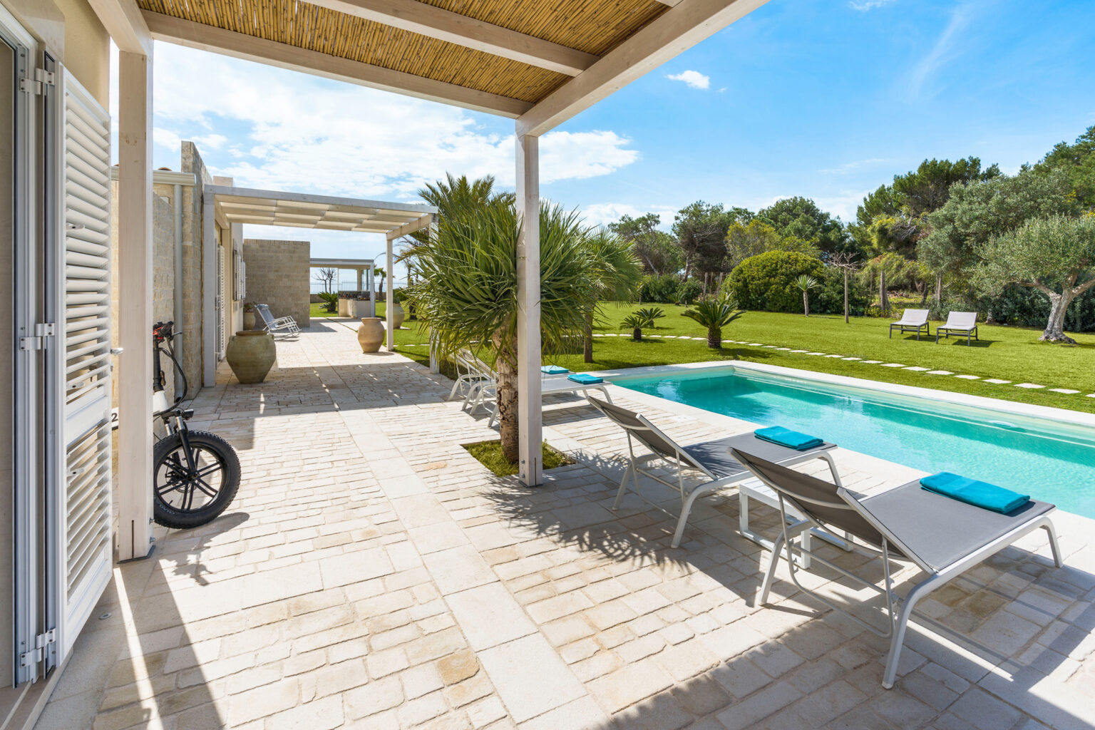 Modern outdoor space featuring a swimming pool, sun loungers with blue towels, and lush green landscaping. A bicycle leans against the wall, and a covered patio provides shade, creating a perfect setting for relaxation and leisure. | Only on SopranoVillas