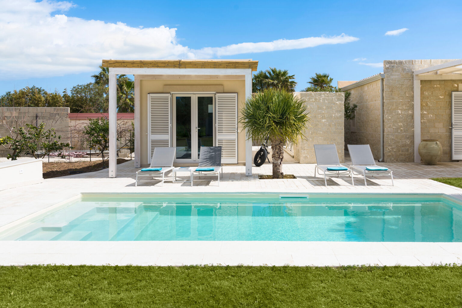 Modern poolside villa with lounge chairs, surrounded by lush greenery and a clear blue sky. The image features a tranquil swimming pool, a stone house with large windows, and a patio area, perfect for relaxation and outdoor living. | Only on SopranoVillas