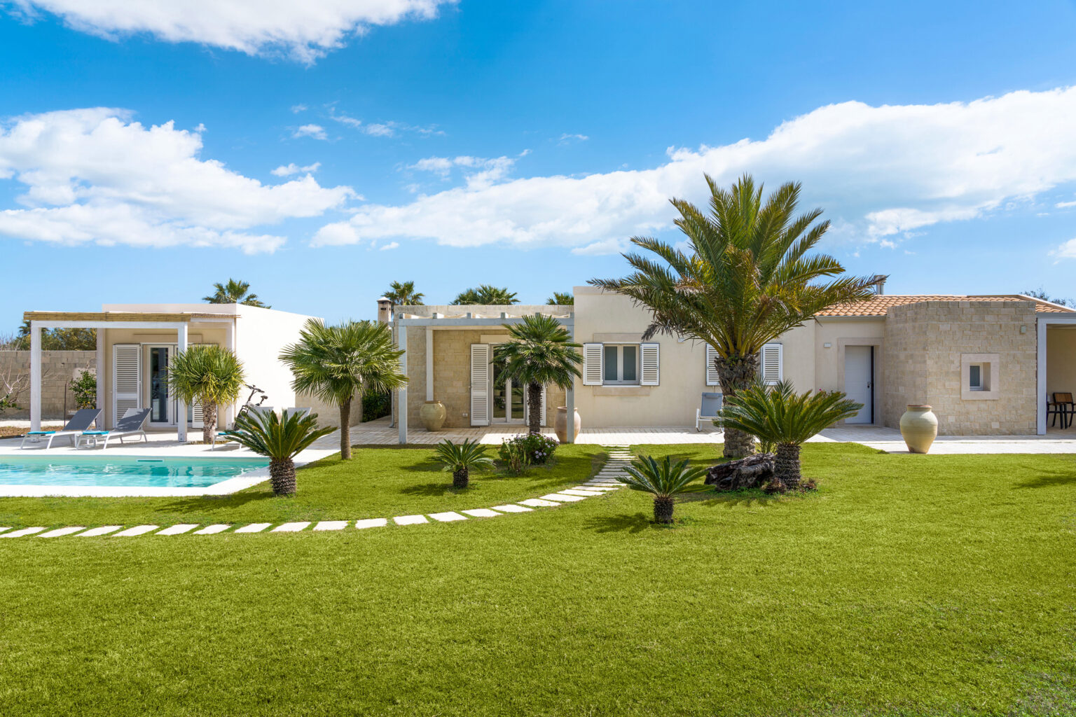 Modern villa with a swimming pool surrounded by lush greenery and palm trees under a clear blue sky. The well-maintained lawn features a stone pathway leading to the house, showcasing a tranquil outdoor living space. | Only on SopranoVillas