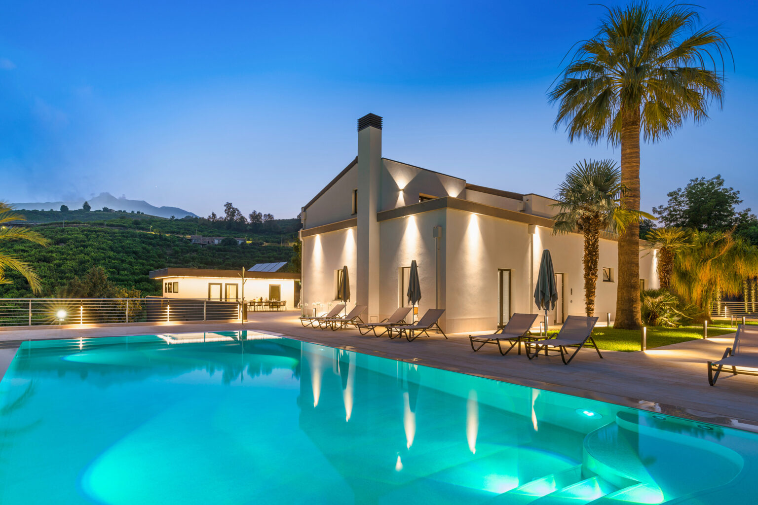 Modern villa with a swimming pool, surrounded by lush greenery and palm trees, illuminated at dusk. The architectural design features large windows, and cozy lounge chairs are positioned around the pool area, offering a serene outdoor retreat. | Only on SopranoVillas