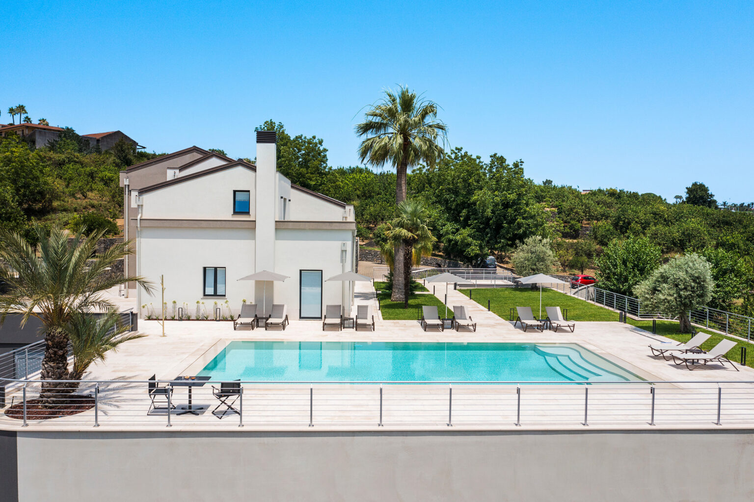 Modern villa with a swimming pool surrounded by palm trees and lush greenery, featuring lounge chairs and a clear blue sky in the background. Ideal for luxury vacation rentals. | Only on SopranoVillas