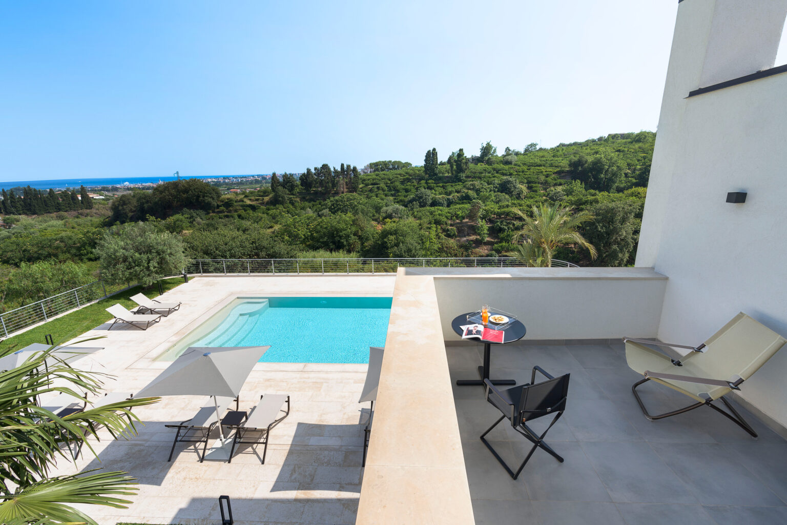 Modern villa terrace with swimming pool, lounge chairs, and scenic views of lush greenery and the ocean. | Only on SopranoVillas