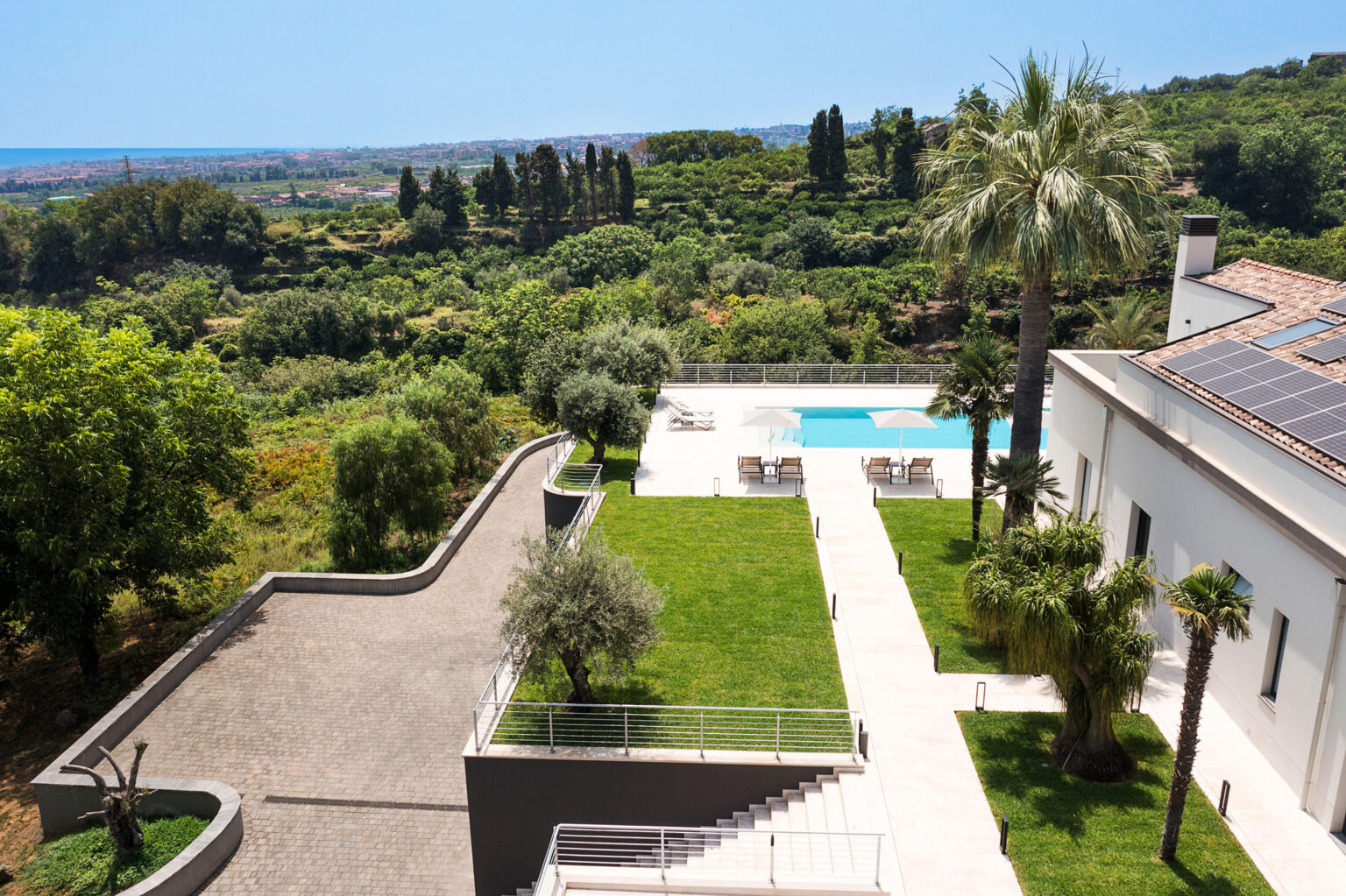 Modern villa with a swimming pool surrounded by lush greenery and palm trees, overlooking a scenic landscape and ocean view. | Only on SopranoVillas