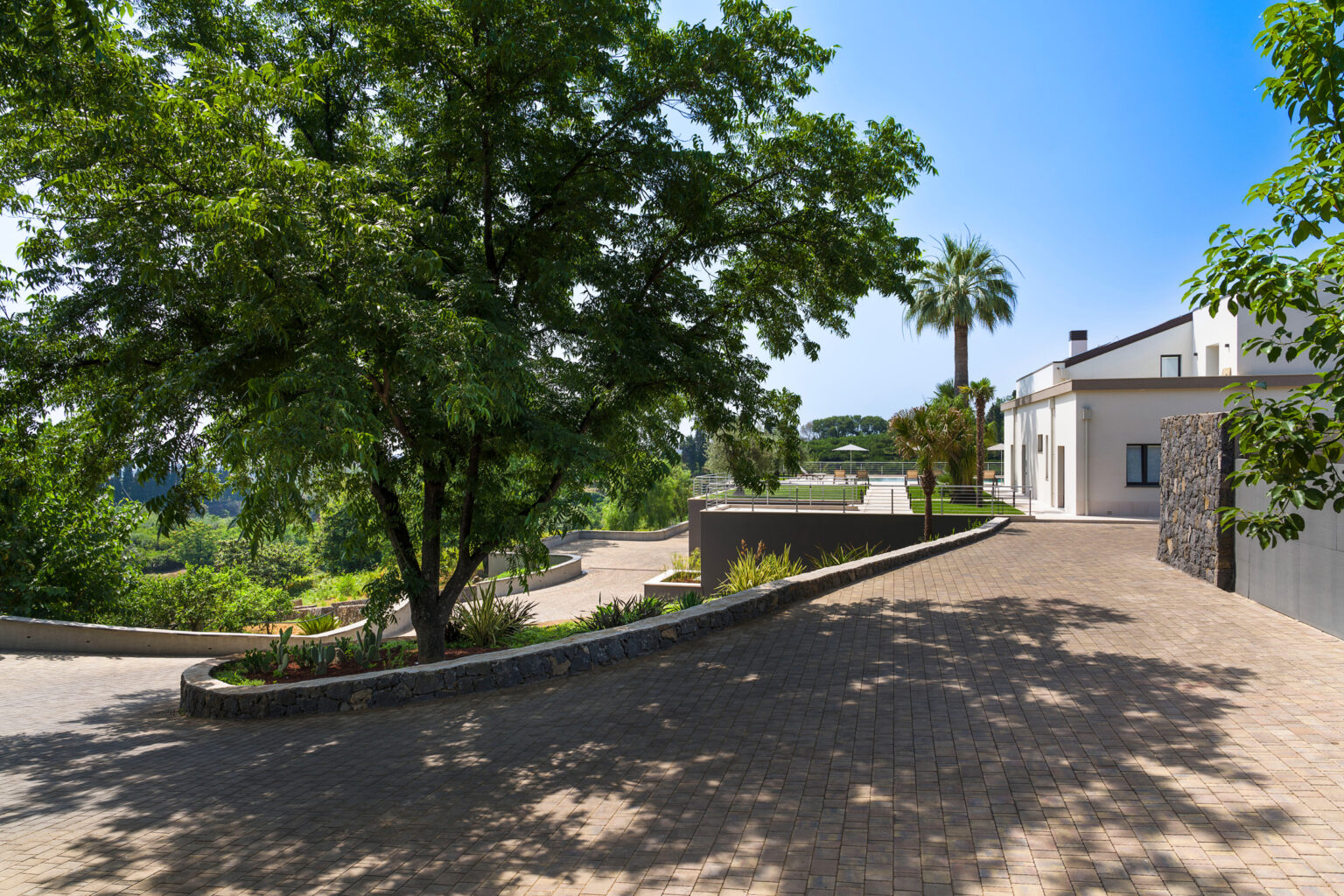 Scenic view of a landscaped pathway lined with lush trees and modern architecture under a clear blue sky, showcasing a tranquil outdoor environment ideal for relaxation and leisure. | Only on SopranoVillas