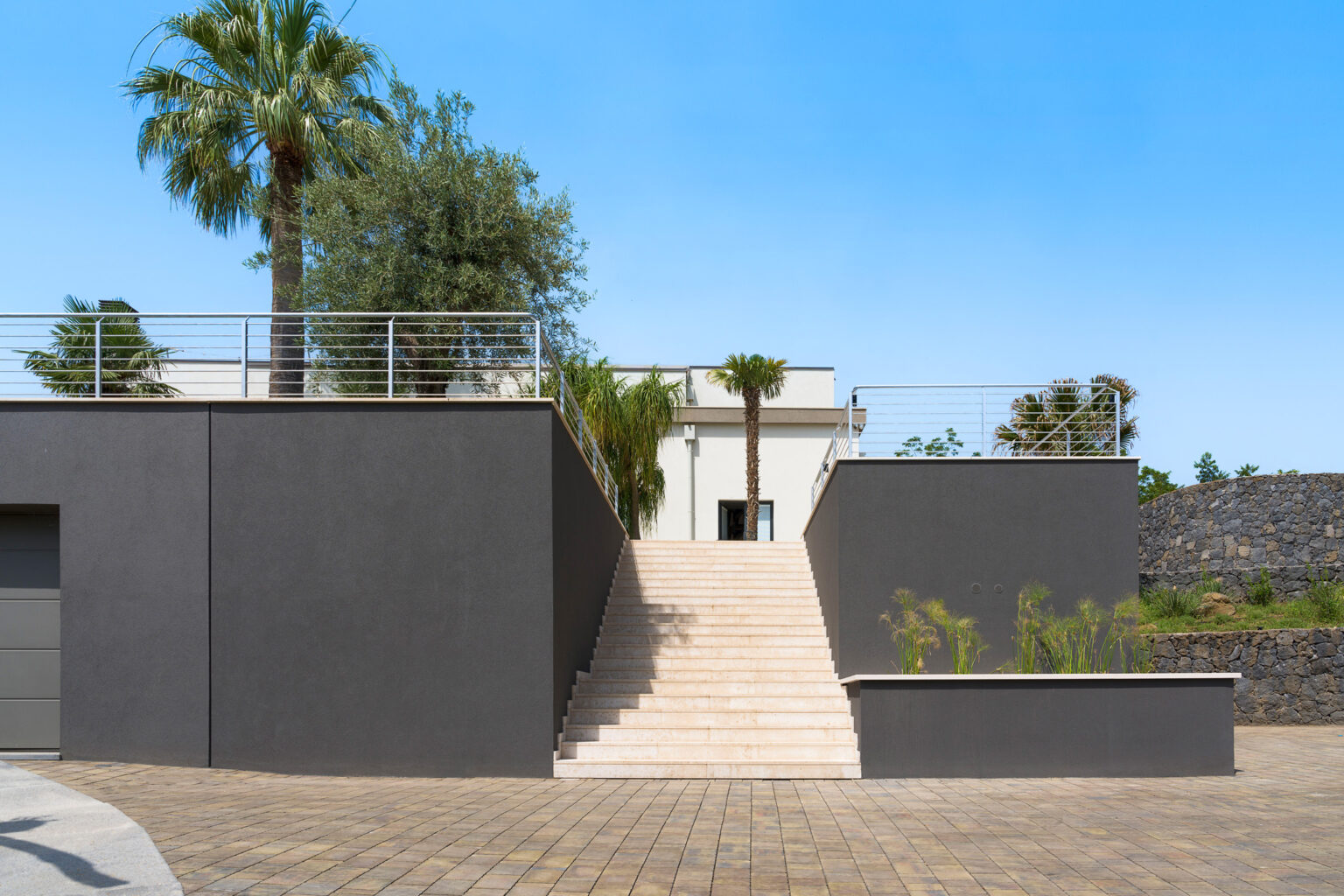 Modern architectural design featuring a sleek gray exterior, stone steps leading to a raised terrace, and lush palm trees against a clear blue sky. Ideal for showcasing contemporary home aesthetics and outdoor living spaces. | Only on SopranoVillas
