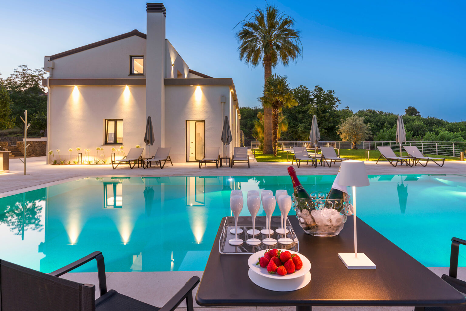 Luxurious villa with a swimming pool at dusk, featuring a table set with champagne glasses and strawberries, surrounded by lounge chairs and palm trees. Ideal for relaxation and upscale outdoor gatherings. | Only on SopranoVillas