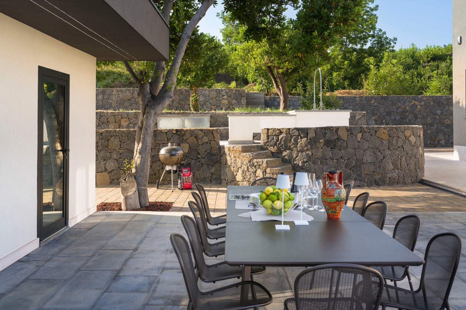 Outdoor dining area featuring a modern table set with fruit, surrounded by lush greenery and stone walls, with a grill in the background, ideal for entertaining guests. | Only on SopranoVillas
